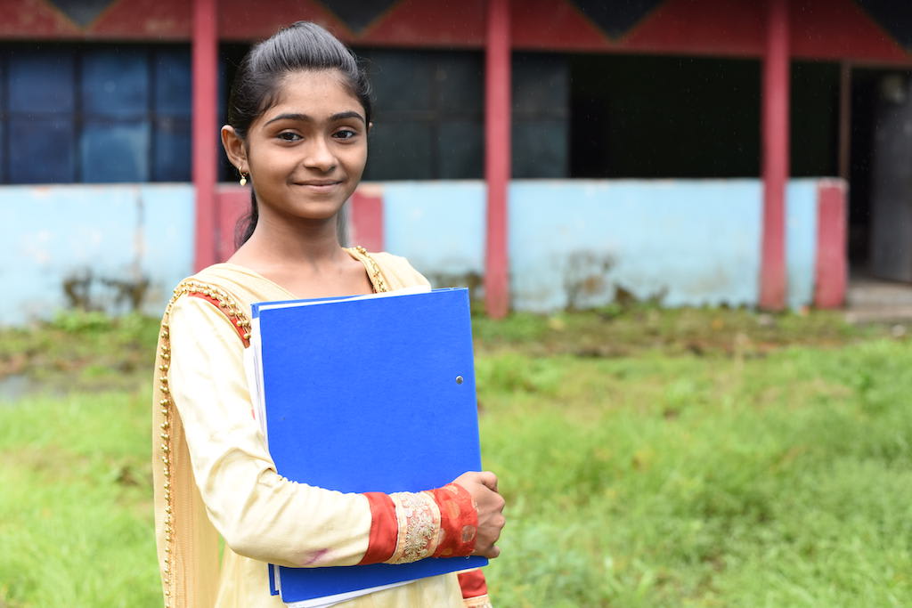Teenage girl wearing yellow and red dress smiles in front of her Compassion centre, holding her workbook.